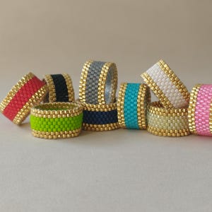 Könnte beinhalten: Eine Sammlung von Perlenringen in verschiedenen Farben, darunter Rot, Schwarz, Grau, Marineblau, Türkis, Weiß, Hellgelb, Grün und Rosa. Jeder Ring hat einen goldfarbenen Rand. Die Ringe sind auf einer neutralen Oberfläche gestapelt.