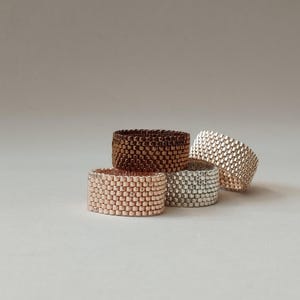 Op de afbeelding: Vier stapelbare ringen gemaakt van geweven kralen. De ringen zijn in tinten bruin, rosé goud en zilver.