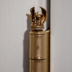 May include: A gold-colored door hinge with a decorative gargoyle finial. The gargoyle is sculpted in a standing position with wings outstretched.