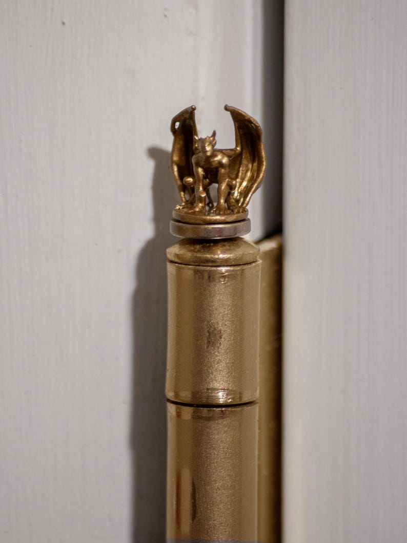 May include: A gold-colored door hinge with a decorative gargoyle finial. The gargoyle is sculpted in a standing position with wings outstretched.