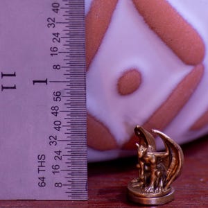 May include: A small, bronze-colored gargoyle figurine with wings, standing on a round base. The figurine is about 1 inch tall and is being measured with a ruler.