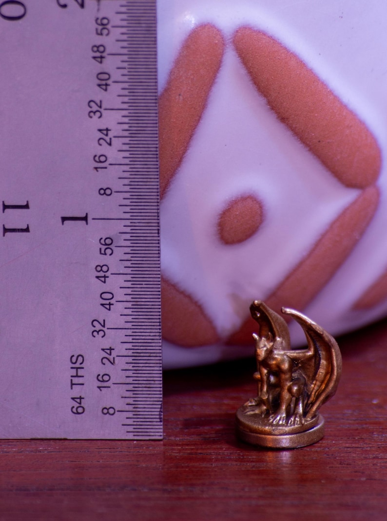 May include: A small, bronze-colored gargoyle figurine with wings, standing on a round base. The figurine is about 1 inch tall and is being measured with a ruler.