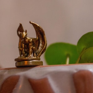 以下が含まれることがあります： 丸い台座の上に立っている、翼のあるガーゴイルの金色の金属製フィギュア。フィギュアは、緑の葉が背景にある白い表面の上に置かれています。