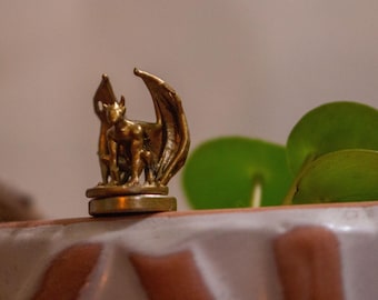 May include: A gold-colored metal figurine of a gargoyle with wings, standing on a round base. The figurine is on a white surface with green leaves in the background.