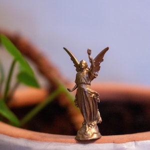 May include: A gold-colored angel figurine with wings outstretched, standing on a small base. The angel is holding a small object in its right hand.