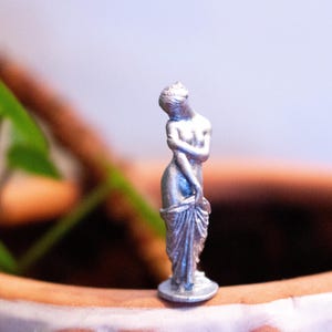 May include: A silver-colored figurine of a woman standing with her arms crossed and a drape around her waist. The figurine is on a small round base.