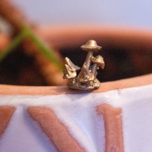 Pode incluir: Uma pequena figura de cogumelo dourada com vários chapéus de cogumelo, sentada na borda de um vaso de terracota.