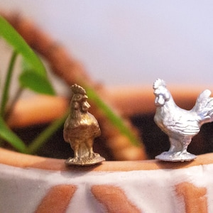 May include: Two small metal rooster figurines, one gold and one silver, are perched on the edge of a terracotta pot with a green plant growing inside.