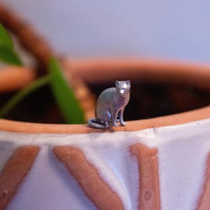 May include: A small, silver-colored cat figurine sits on the rim of a terracotta-colored ceramic pot. The cat is in a seated position, with its tail curled around its body. The pot has a white and brown patterned design.