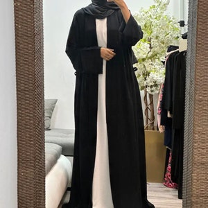Op de afbeelding: Een zwarte abaya met lange mouwen en een vloeiende silhouet. De abaya is open aan de voorkant en heeft een wit onderhemd.