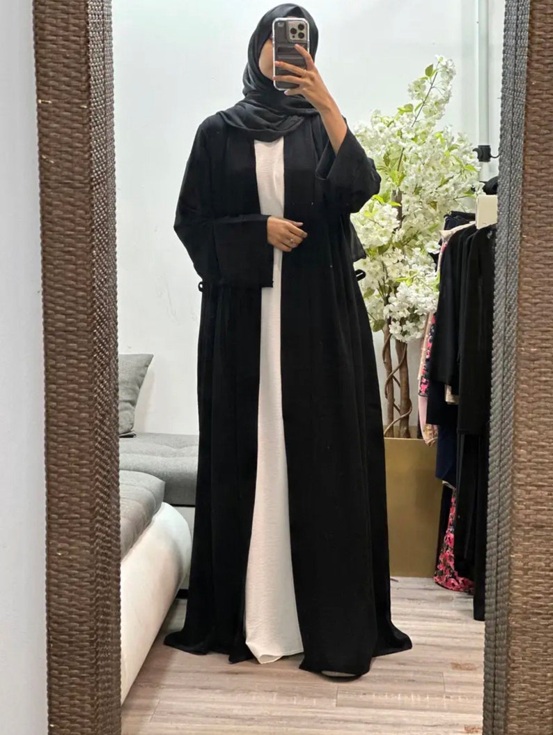 Op de afbeelding: Een zwarte abaya met lange mouwen en een vloeiende silhouet. De abaya is open aan de voorkant en heeft een wit onderhemd.