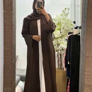Op de afbeelding: Een bruine abaya met lange mouwen en een vloeiende silhouet. De abaya wordt gedragen over een wit onderhemd.