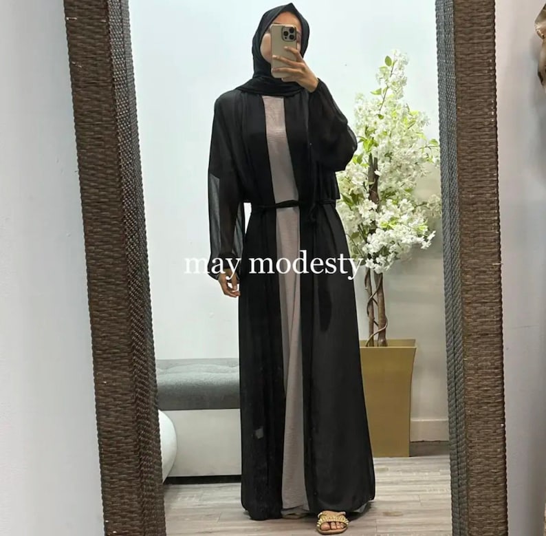 May include: A black sheer long-sleeved cardigan with a tie belt. The cardigan is worn over a long-sleeved dress. The text "may modesty" is visible on the bottom of the image.