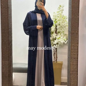 May include: A navy blue long-sleeved abaya with a long, flowing design. The abaya is worn over a light gray dress. The text "may modesty" is visible on the image.