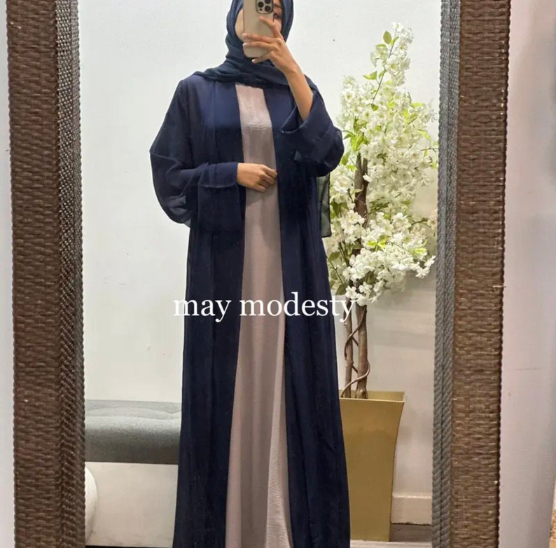 May include: A navy blue long-sleeved abaya with a long, flowing design. The abaya is worn over a light gray dress. The text "may modesty" is visible on the image.