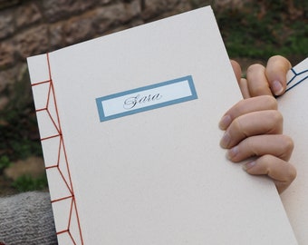 Puede incluir: Un cuaderno color crema con el nombre "Sara" en una etiqueta azul claro. La encuadernación presenta un diseño geométrico cosido con hilo naranja. Otro cuaderno es parcialmente visible.