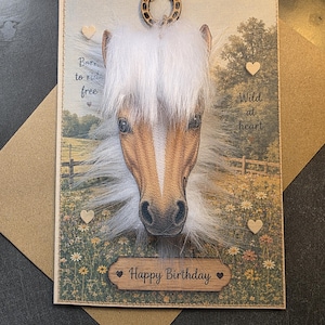 May include: A rectangular greeting card featuring a 3D horse head with white faux fur mane, a wooden horseshoe accent, and small wooden hearts. The background shows a field with flowers and text reading Born to ride free, Wild at heart, and Happy Birthday.