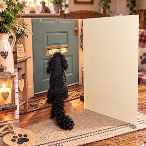 May include: A greeting card featuring a black curly-haired dog sitting and looking through a mail slot in a sage green door. The card is open, showing a blank cream-colored interior panel. The front of the card displays a detailed scene of a dog waiting at a doorway, with a wooden heart ornament with a paw print design placed on the table in the foreground.