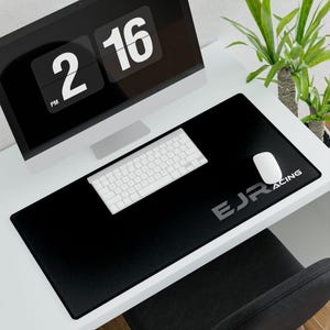 Può includere: Un tappetino da scrivania rettangolare nero con la scritta "EJR RACING" in argento. Una tastiera e un mouse bianchi sono sul tappetino. Un monitor di computer mostra l'ora: 14:16. Una pianta verde è sullo sfondo.