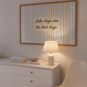 K&ouml;nnte beinhalten: Ein rechteckiges Wandbild in einem hellen Holzrahmen mit vertikalen hellblauen und wei&szlig;en Streifen und einem cremefarbenen Rechteck in der Mitte, das den schwarzen kursiven Text Lake days are the best days &uuml;ber einem wei&szlig;en Schrank zeigt.