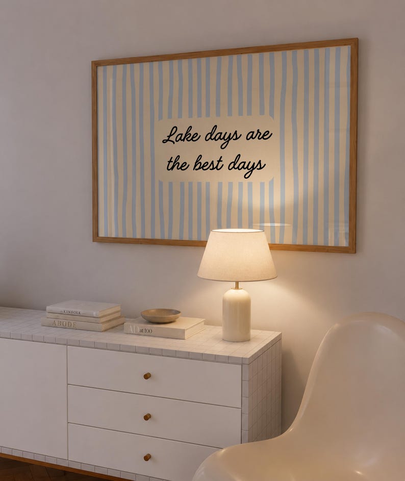 K&ouml;nnte beinhalten: Ein rechteckiges Wandbild in einem hellen Holzrahmen mit vertikalen hellblauen und wei&szlig;en Streifen und einem cremefarbenen Rechteck in der Mitte, das den schwarzen kursiven Text Lake days are the best days &uuml;ber einem wei&szlig;en Schrank zeigt.