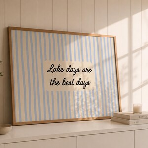K&ouml;nnte beinhalten: Ein rechteckiger gerahmter Wandkunstdruck mit einem vertikalen Streifenmuster in Hellblau und Creme. In der Mitte befindet sich ein cremefarbener rechteckiger Block mit dem schwarzen kursiven Text, Lake days are the best days.