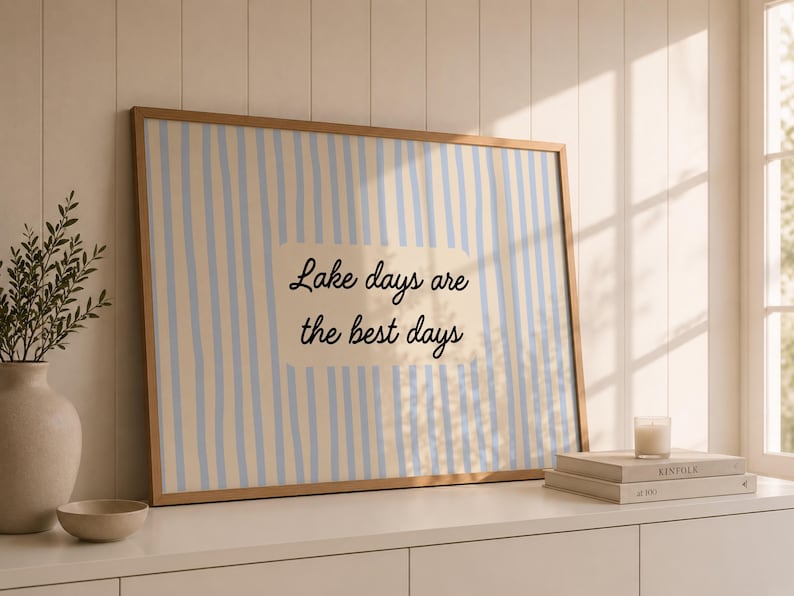 K&ouml;nnte beinhalten: Ein rechteckiger gerahmter Wandkunstdruck mit einem vertikalen Streifenmuster in Hellblau und Creme. In der Mitte befindet sich ein cremefarbener rechteckiger Block mit dem schwarzen kursiven Text, Lake days are the best days.
