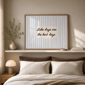 K&ouml;nnte beinhalten: Ein rechteckiger Wandkunstdruck in einem Holzrahmen mit einem vertikalen Streifenmuster in Hellblau und Creme. In der Mitte befindet sich ein cremefarbenes rechteckiges Etikett mit dem schwarzen Text "Lake days are the best days" in Schreibschrift.