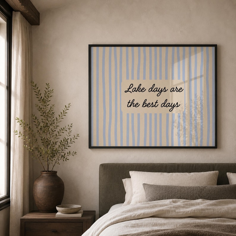 K&ouml;nnte beinhalten: Ein rechteckiger Wandkunstdruck in einem schwarzen Rahmen mit einem vertikalen Streifenmuster in Hellblau und Beige. Zentriert auf dem Druck befindet sich ein beiger rechteckiger Block mit dem schwarzen kursiven Text, Lake days are the best days.