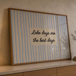 K&ouml;nnte beinhalten: Ein rechteckiges gerahmtes Wandbild mit einem vertikalen Streifenmuster in Hellblau und Creme. In der Mitte befindet sich ein cremefarbenes Rechteck mit dem schwarzen kursiven Text: Lake days are the best days.