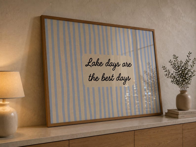 K&ouml;nnte beinhalten: Ein rechteckiges gerahmtes Wandbild mit einem vertikalen Streifenmuster in Hellblau und Creme. In der Mitte befindet sich ein cremefarbenes Rechteck mit dem schwarzen kursiven Text: Lake days are the best days.