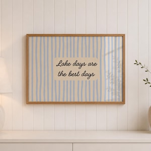 K&ouml;nnte beinhalten: Ein rechteckiger Wandkunstdruck in einem Holzrahmen mit einem vertikalen Streifenmuster in Hellblau und Beige. In der Mitte befindet sich ein beiger rechteckiger Block mit dem schwarzen kursiven Text, Lake days are the best days.