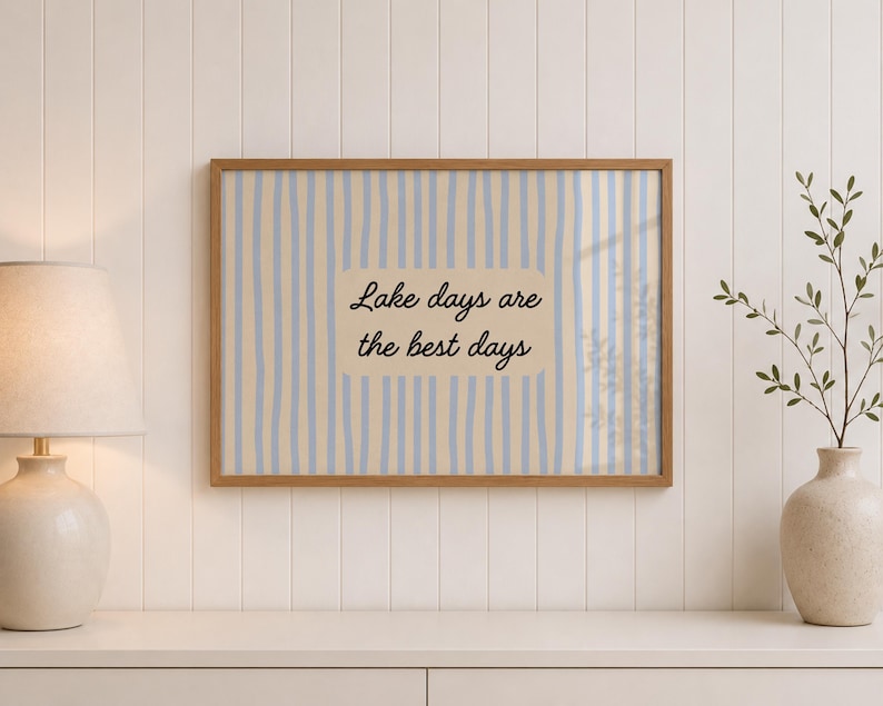 K&ouml;nnte beinhalten: Ein rechteckiger Wandkunstdruck in einem Holzrahmen mit einem vertikalen Streifenmuster in Hellblau und Beige. In der Mitte befindet sich ein beiger rechteckiger Block mit dem schwarzen kursiven Text, Lake days are the best days.