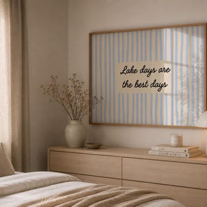 K&ouml;nnte beinhalten: Ein rechteckiger Wandkunstdruck in einem hellen Holzrahmen mit vertikalen hellblauen und beigen Streifen und dem schwarzen Text Lake days are the best days, der in einem Rechteck auf dem Druck zentriert ist.