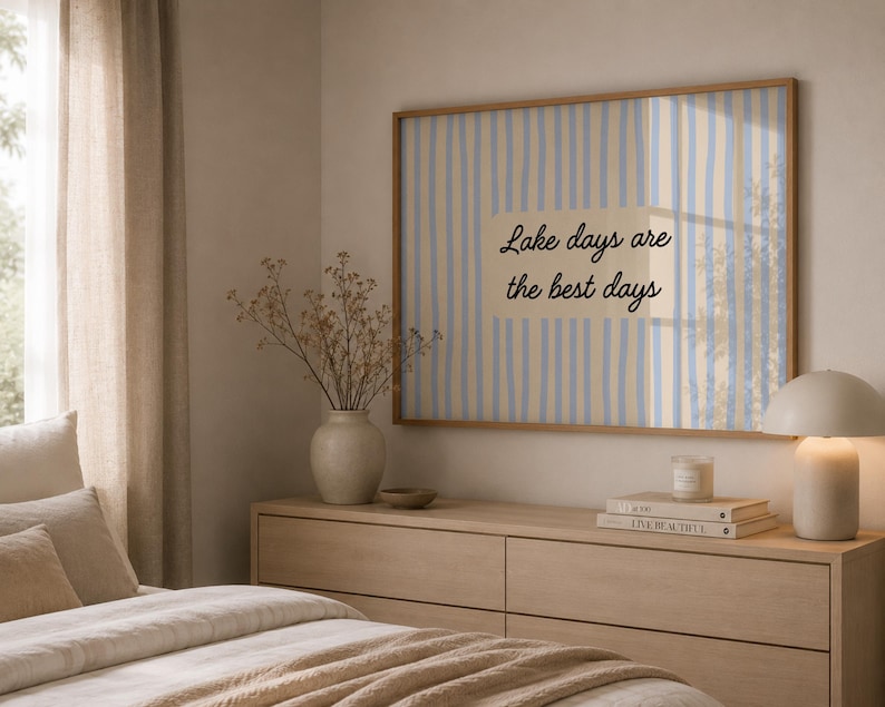 K&ouml;nnte beinhalten: Ein rechteckiger Wandkunstdruck in einem hellen Holzrahmen mit vertikalen hellblauen und beigen Streifen und dem schwarzen Text Lake days are the best days, der in einem Rechteck auf dem Druck zentriert ist.