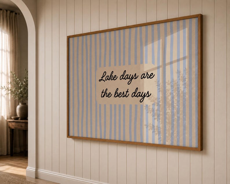 May include: A rectangular wall art piece in a wooden frame featuring a light beige background with vertical blue stripes. The center displays the black cursive text "Lake days are the best days" printed on a solid, lighter beige rectangular patch.