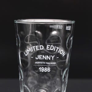 Könnte beinhalten: Klares Glas mit strukturiertem Muster und dem weißen Schriftzug "LIMITED EDITION - JENNY - PERFEKTE MISCHUNG 1986". Das zylindrische Glas ist leer, vor schwarzem Hintergrund.