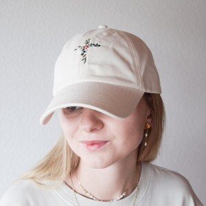 Könnte beinhalten: Cremefarbene Baseballkappe mit gebogenem Schirm. Die Kappe hat ein gesticktes Blumendesign in Grün-, Rot- und Brauntönen auf der Vorderseite. Der Hut ist aus weichem Stoff und hat ein klassisches Sechs-Panel-Design.