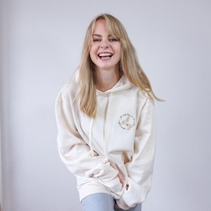 Könnte beinhalten: Cremefarbener Kapuzenpullover mit Kordelzug und einem kleinen gestickten Design auf der linken Brust. Das Design zeigt ein Kaninchen in einem floralen Kranz. Der Hoodie hat eine lockere Passform und lange Ärmel.