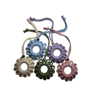 Puede incluir: Seis adornos florales de macramé hechos a mano en varios colores, incluyendo azul claro, lavanda y verde oliva. Cada flor tiene un agujero central y un cordón para colgar con una cuenta. Los adornos se muestran sobre un fondo blanco.