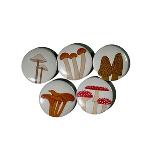 May include: Set of six mushroom-themed button pins. The pins feature different types of mushrooms, including white mushrooms, brown mushrooms, and red and white mushrooms.