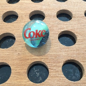 May include: A blue glass marble with a red "Coke" logo and the words "Marca Reg" on a wooden board with circular holes.