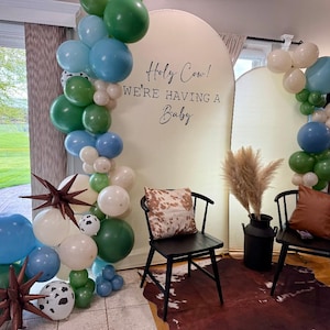 May include: A baby shower backdrop featuring a cream-colored arch with the text Holy Cow! WE'RE HAVING A BABY, decorated with blue, green, and white balloons, brown star-shaped foil balloons, two black chairs, and a cow-print rug.