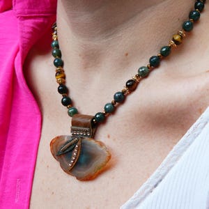 Può includere: Una collana artigianale con un ciondolo in agata nei toni del blu, marrone e arancione. Il ciondolo ha una forma irregolare ed è impreziosito da metallo e piccoli cristalli. La catena di perline ha perline verdi, marroni e dorate.