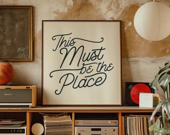May include: A white framed print with black calligraphy that reads "This Must Be The Place" hanging on a wall above a wooden bookshelf filled with records and books. The wall is a light beige color with a textured finish.