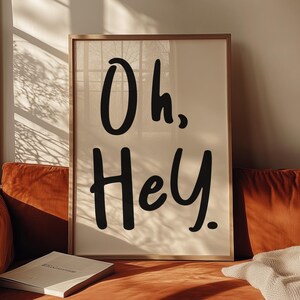 May include: A framed print with the text "Oh, Hey" in black lettering on a white background. The print is hanging on a wall above a brown couch.