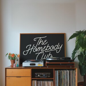 May include: A wooden record player cabinet with two turntables, a cassette player, and shelves filled with vinyl records. A black sign above the cabinet reads "The Homebody Club".