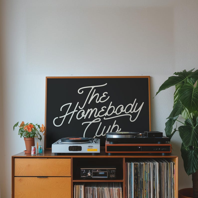 May include: A wooden record player cabinet with two turntables, a cassette player, and shelves filled with vinyl records. A black sign above the cabinet reads "The Homebody Club".