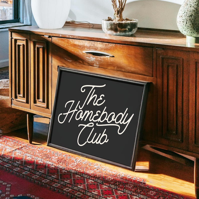 May include: Black framed sign with white text that reads "The Homebody Club" on a wooden surface.