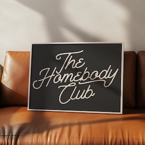 May include: A black framed print with white text that reads "The Homebody Club" in a cursive font.